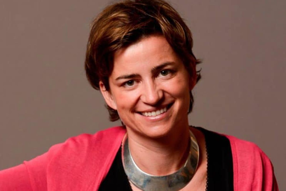 Headshot of Sophie Lambin smiling, looking directly into the camera against a neutral background. She wears a coral cardigan, black top and chunky silver necklace. Her hair is short and brown, with gold highlights,