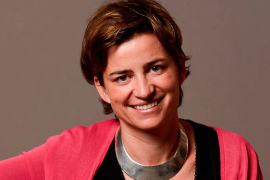 Headshot of Sophie Lambin smiling, looking directly into the camera against a neutral background. She wears a coral cardigan, black top and chunky silver necklace. Her hair is short and brown, with gold highlights,