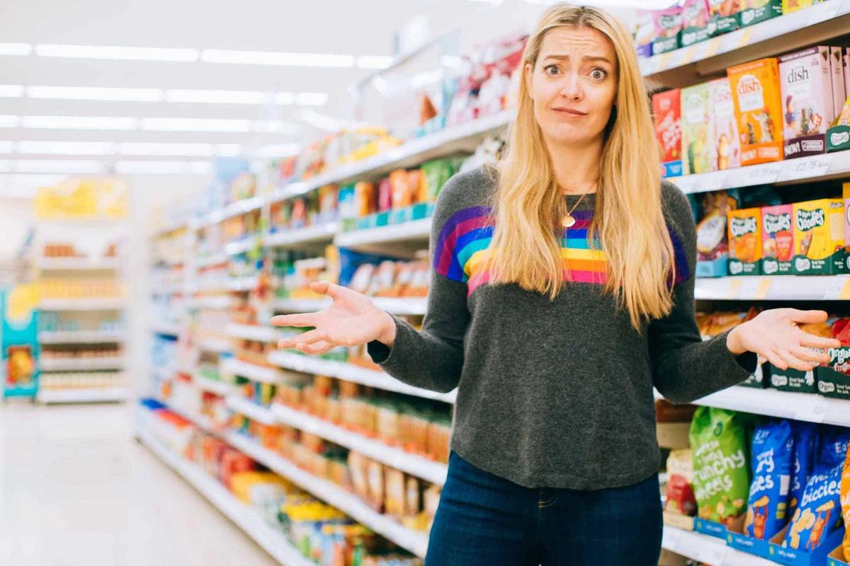 White person with long blonde hair stands in front of a supermarket aisle with a puzzled look on their face.