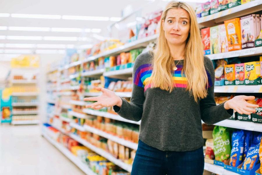 White person with long blonde hair stands in front of a supermarket aisle with a puzzled look on their face.