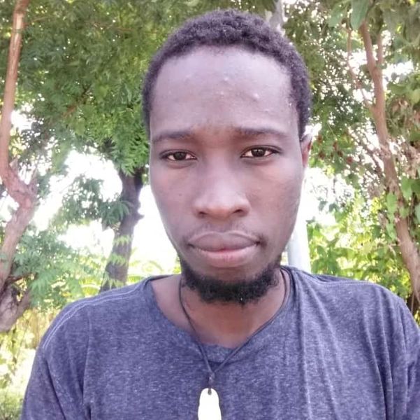A selfie of a black man with a short beard. They're wearing a grey blue top and standing in a forest.