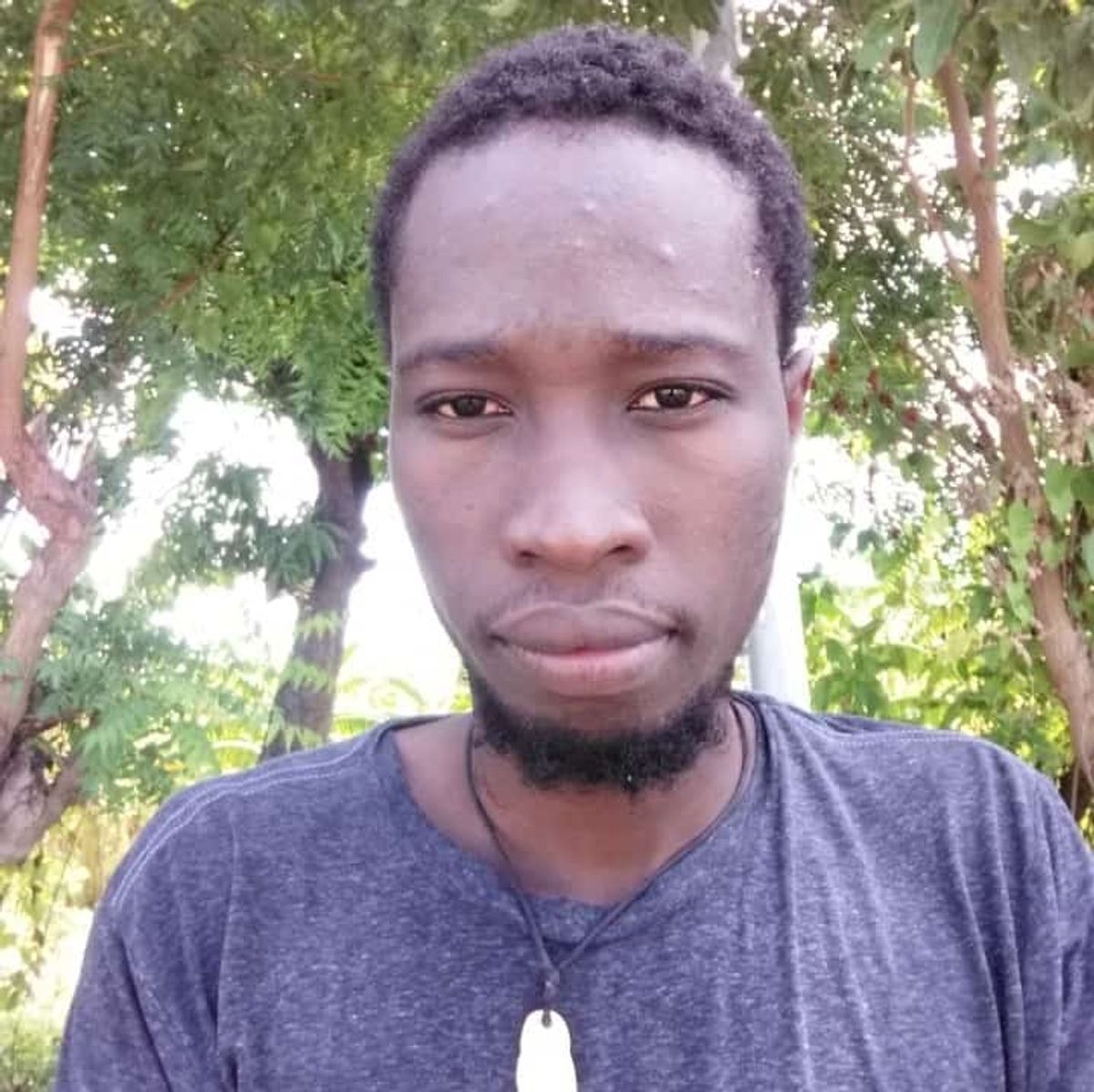 A selfie of a black man with a short beard. They're wearing a grey blue top and standing in a forest.
