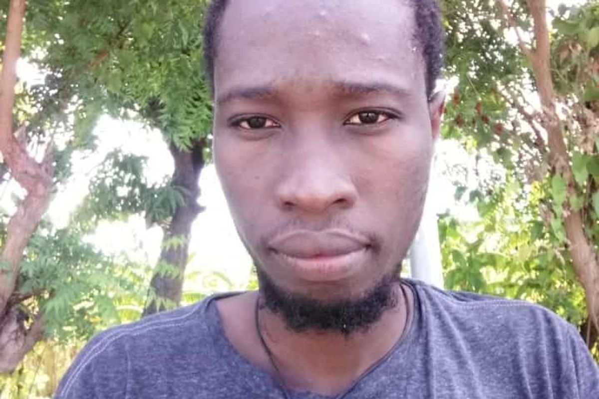 A selfie of a black man with a short beard. They're wearing a grey blue top and standing in a forest.