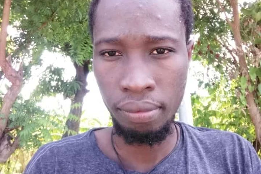 A selfie of a black man with a short beard. They're wearing a grey blue top and standing in a forest.
