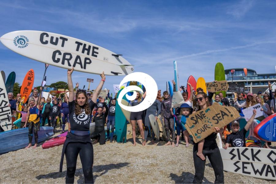 Surfers Against Sewage community campaigners and surfers holding boards with anti-sewage pollution messages at a coastal protest.