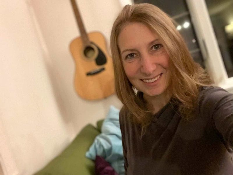 Lara Seaver is smiling, standing in front of a guitar hanging on a wall.