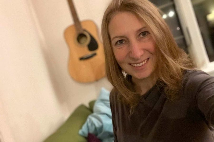 Lara Seaver is smiling, standing in front of a guitar hanging on a wall.