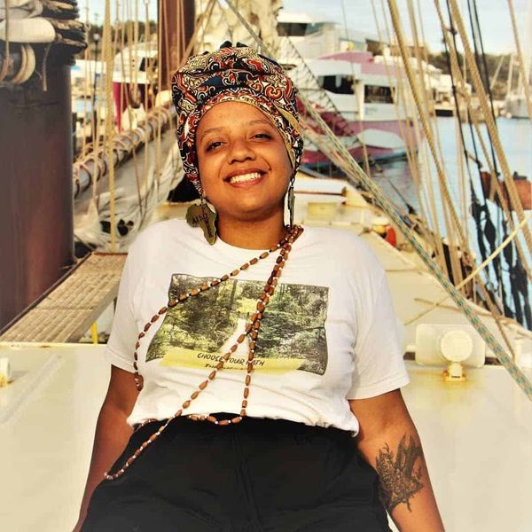 A black woman is sat on the edge of a sail boat wearing a headdress, and wooden jewellery.