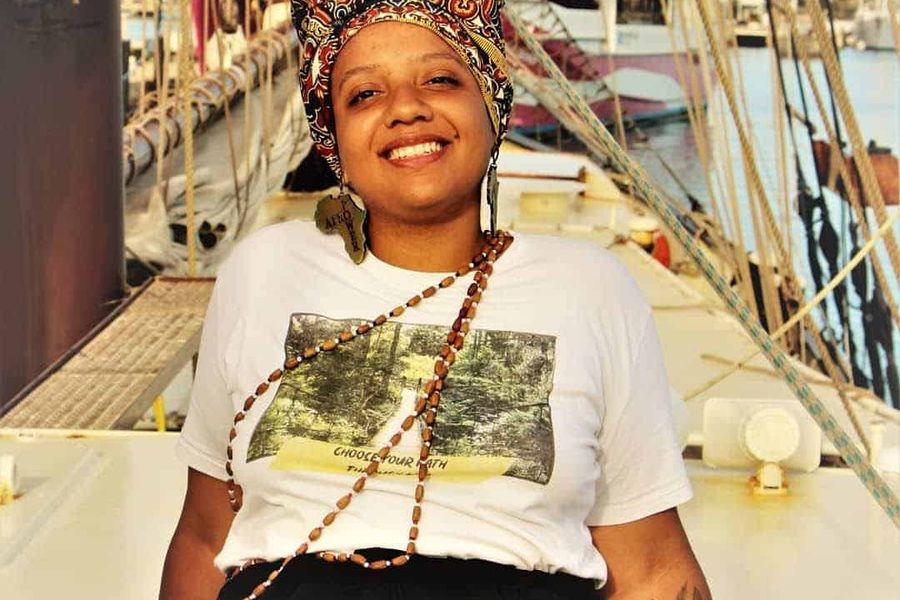 A black woman is sat on the edge of a sail boat wearing a headdress, and wooden jewellery.
