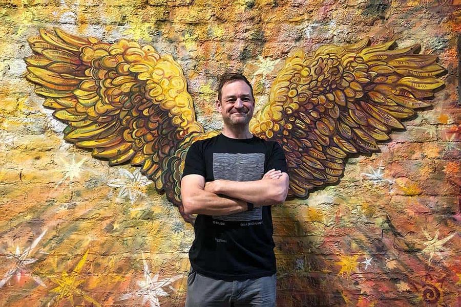 A white man is standing cross-armed wearing a black t-shirt and grey trousers. He is in front of a gold mural of a pair of angel wings.