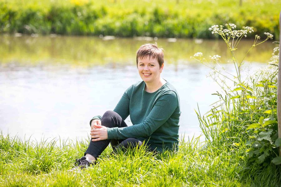 Fiona Stainer from Greenhouse Communications. Fiona is sitting on the bright green grass in front of the River Avon. Behind her you can see the opposite riverbank. Fiona has one leg tucked into her chest which she is holding with her hands in a relaxed pose. She is smiling, wearing a green sweater, black skinny jeans and black trainers.