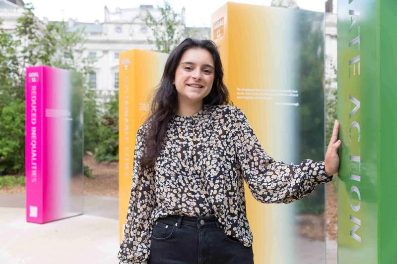 Sindhu has long black hair, and is standing in a circle of pillars that represent the 17 sustainable development goals. She is wearing a floral print long sleeve blouse and black jeans.