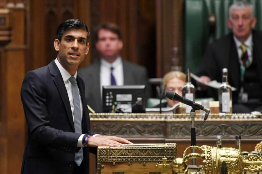 A dark skinned man in a suit stands at a microphone speaking