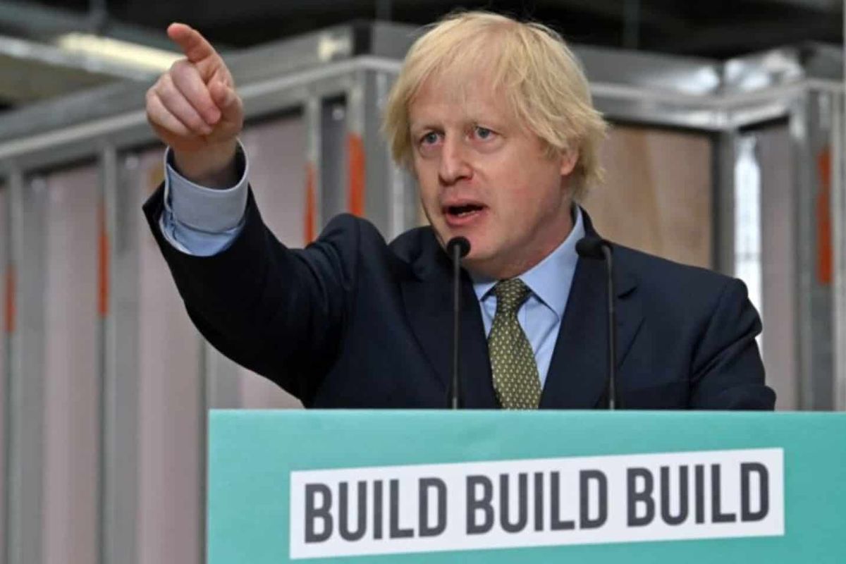 A white man in a suit points his finger, and speaks from behind a stand which has 'Build Build Build' written on the front