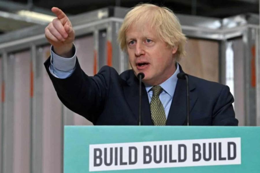 A white man in a suit points his finger, and speaks from behind a stand which has 'Build Build Build' written on the front