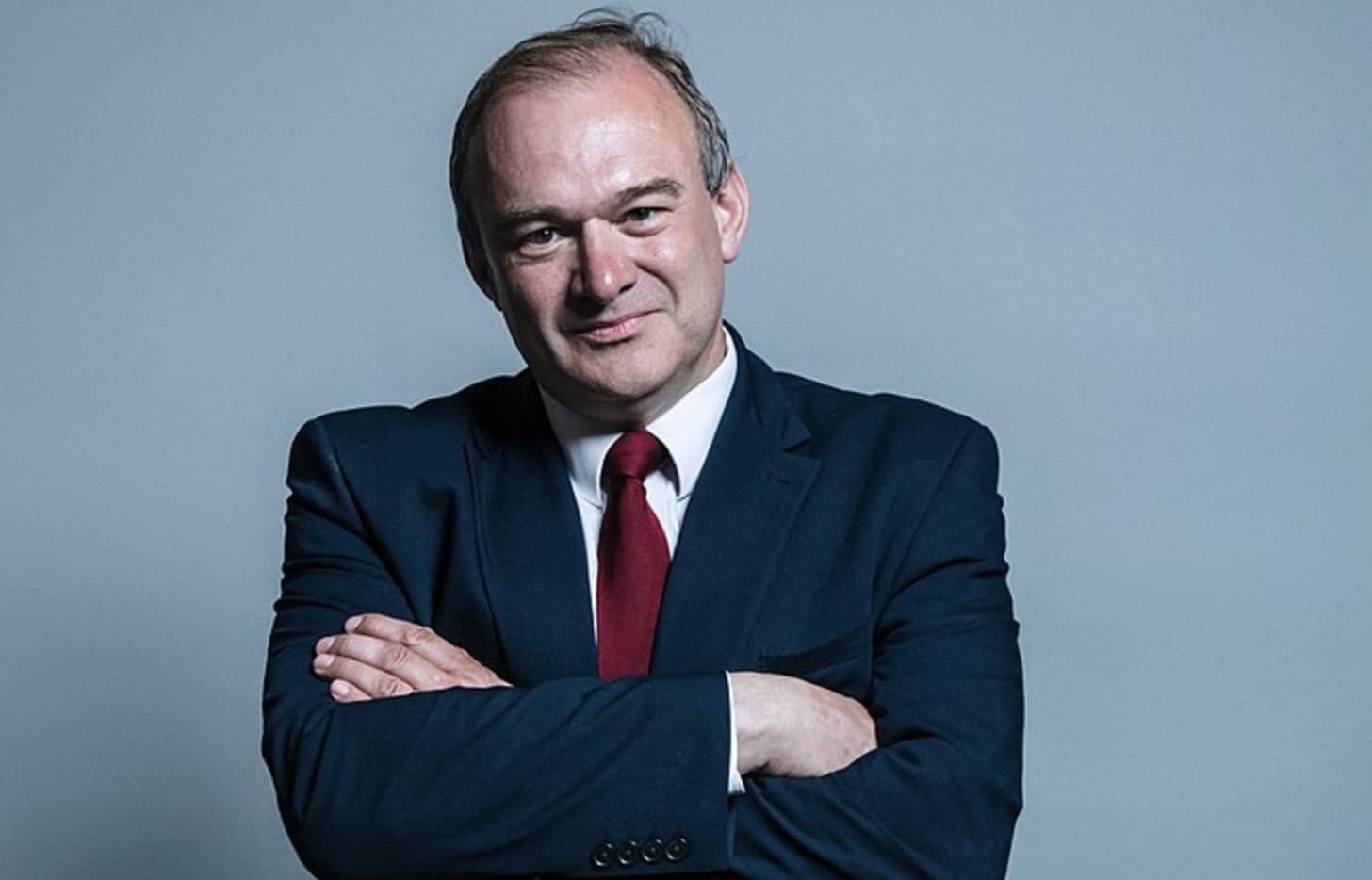 A white man in a suit looks directly at camera, with his arms crossed