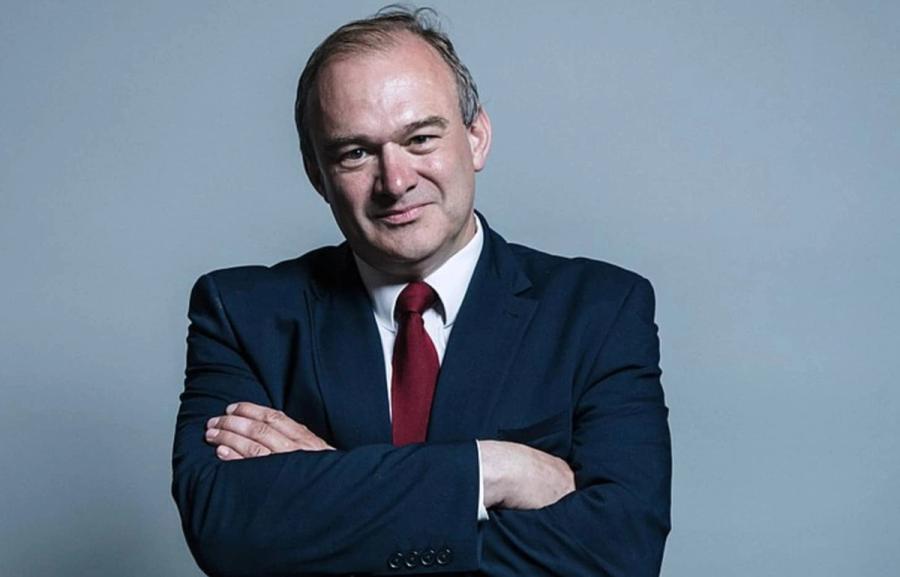 A white man in a suit looks directly at camera, with his arms crossed