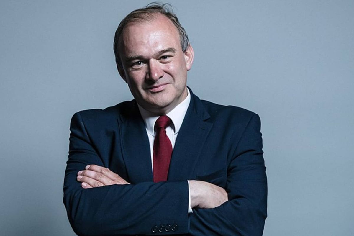 A white man in a suit looks directly at camera, with his arms crossed