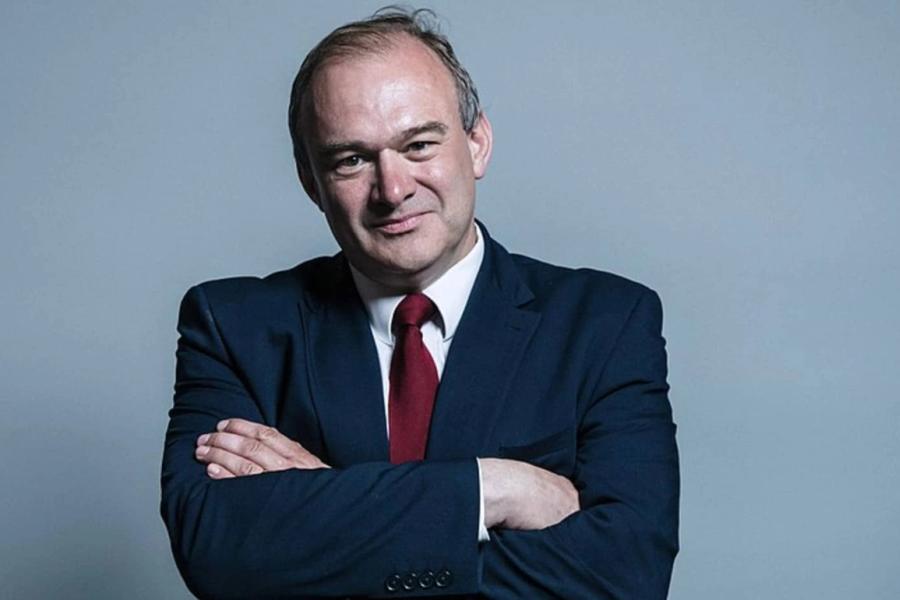 A white man in a suit looks directly at camera, with his arms crossed