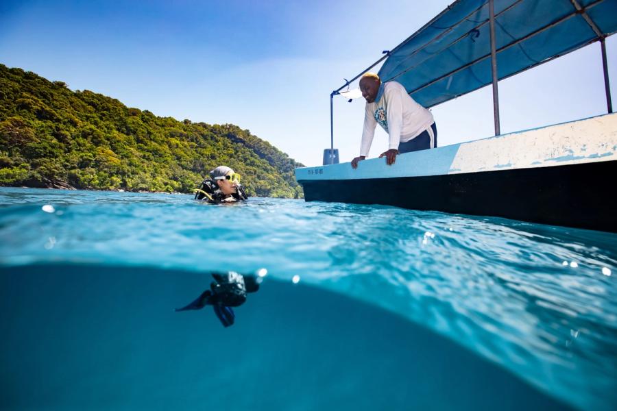 A scuba diver in clear turquoise water beside a boat, a crew member leaning over the side, with a forested coastline behind.