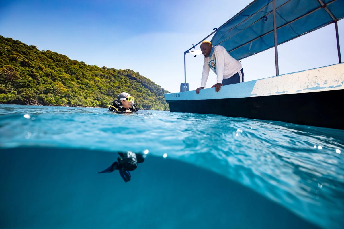 A scuba diver in clear turquoise water beside a boat, a crew member leaning over the side, with a forested coastline behind.