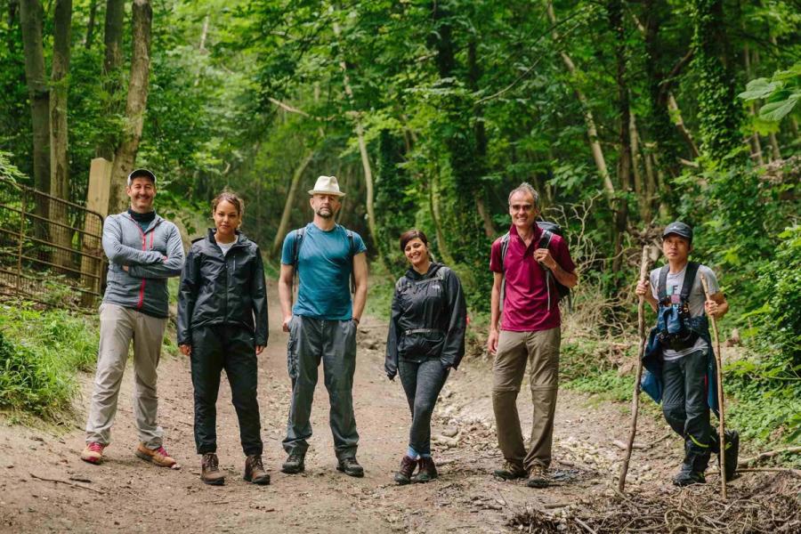 Six people in walking clothes are stood in a forest on a dirt road.