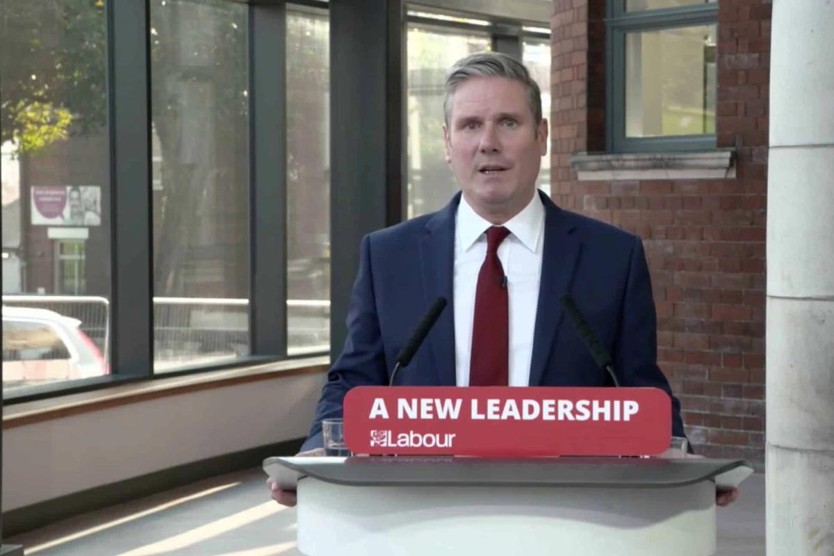 A white man in a suit stood at a press stand where a sign reads ' A New Leadership / Labour'
