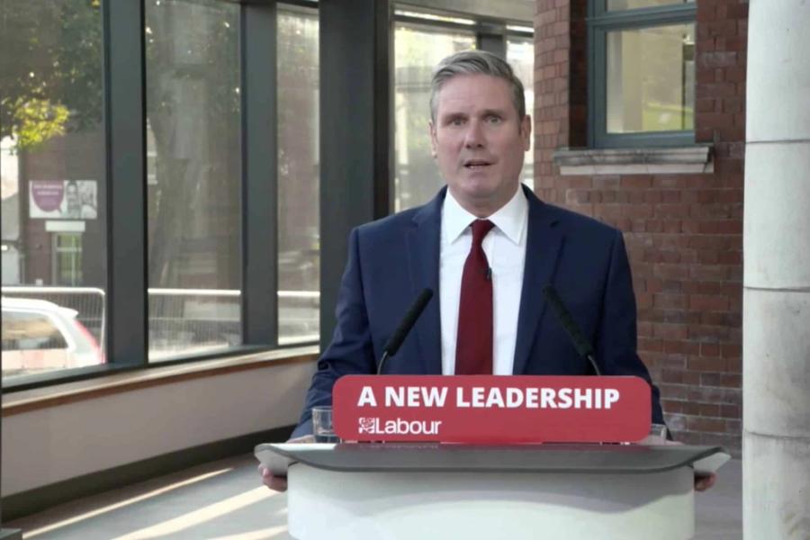 A white man in a suit stood at a press stand where a sign reads ' A New Leadership / Labour'