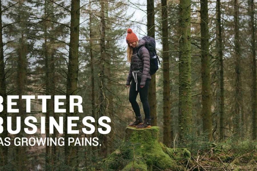 Person stood on a mossy tree stump in a wood. They're wearing Vivobarefoot shoes, a bobble hat and a rucksack. Text reads "Better business has growing pains."