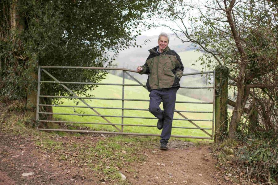 A white man leans against a metal gate in front of a field