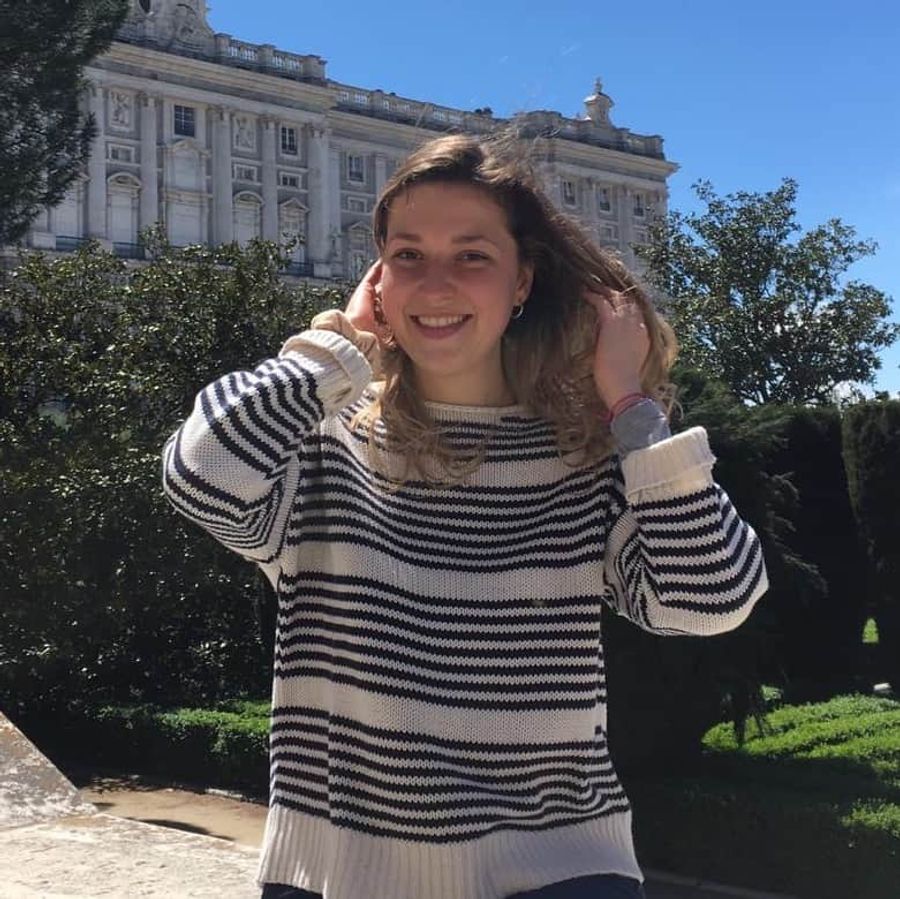 Maria is white with brown hairand is wearing a cream and black stripy jumper and she is stood in front of a large white building.