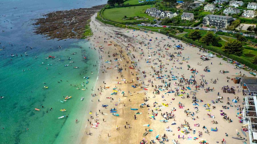Aerial view of protesters gathered on Gyllyngvase Beach in Falmouth ahead of the paddle out. A strip of sandy beach takes up the centre of the image, with aquamarine sea to the left and green fields to the right. The beach is very busy, with many people assembled in swimsuits and wetsuits ready for the paddle out.