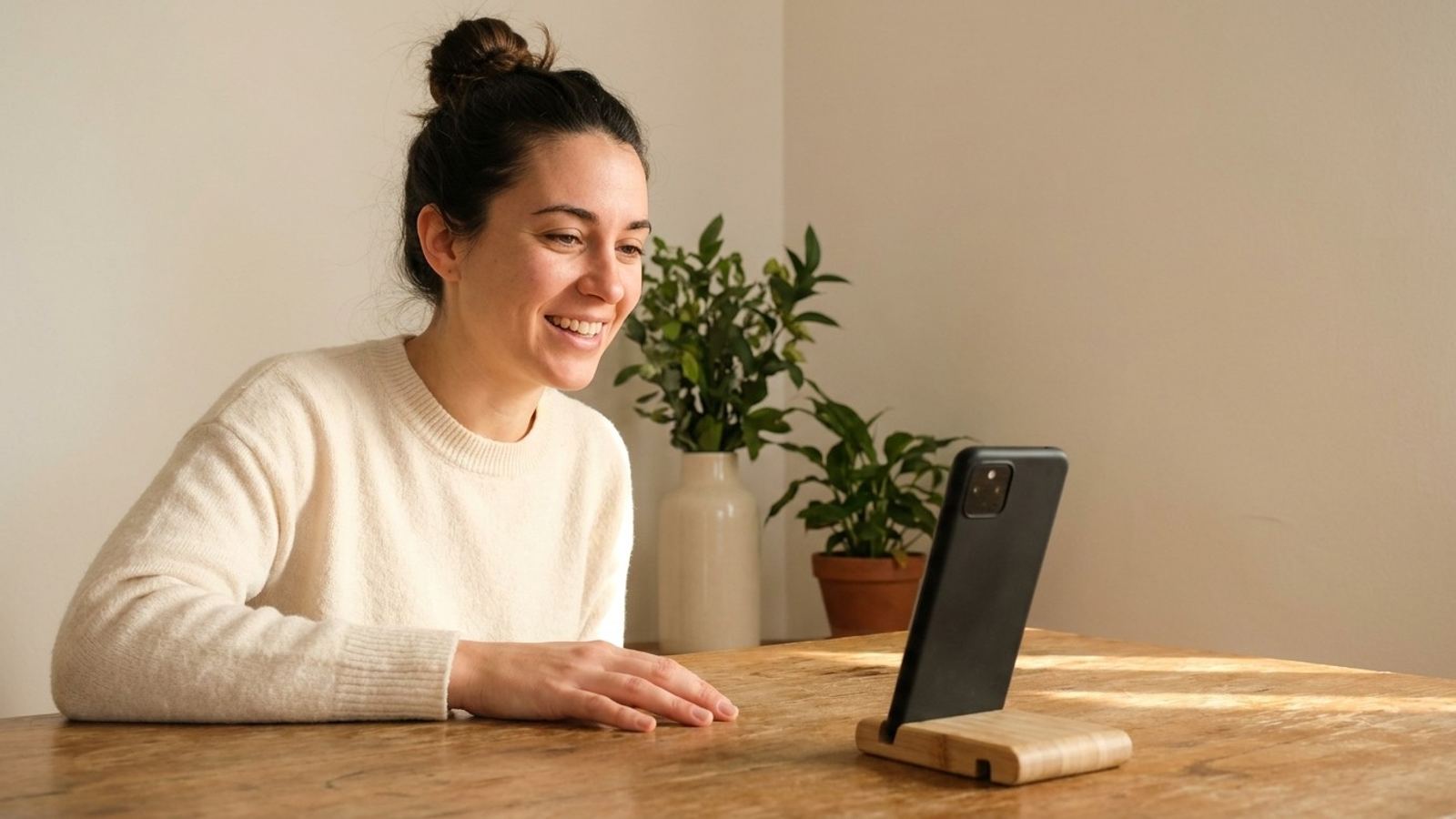 Une jeune femme souriante en pleine conversation, la main posée sur la table, face à son téléphone dressé sur un support en bois.