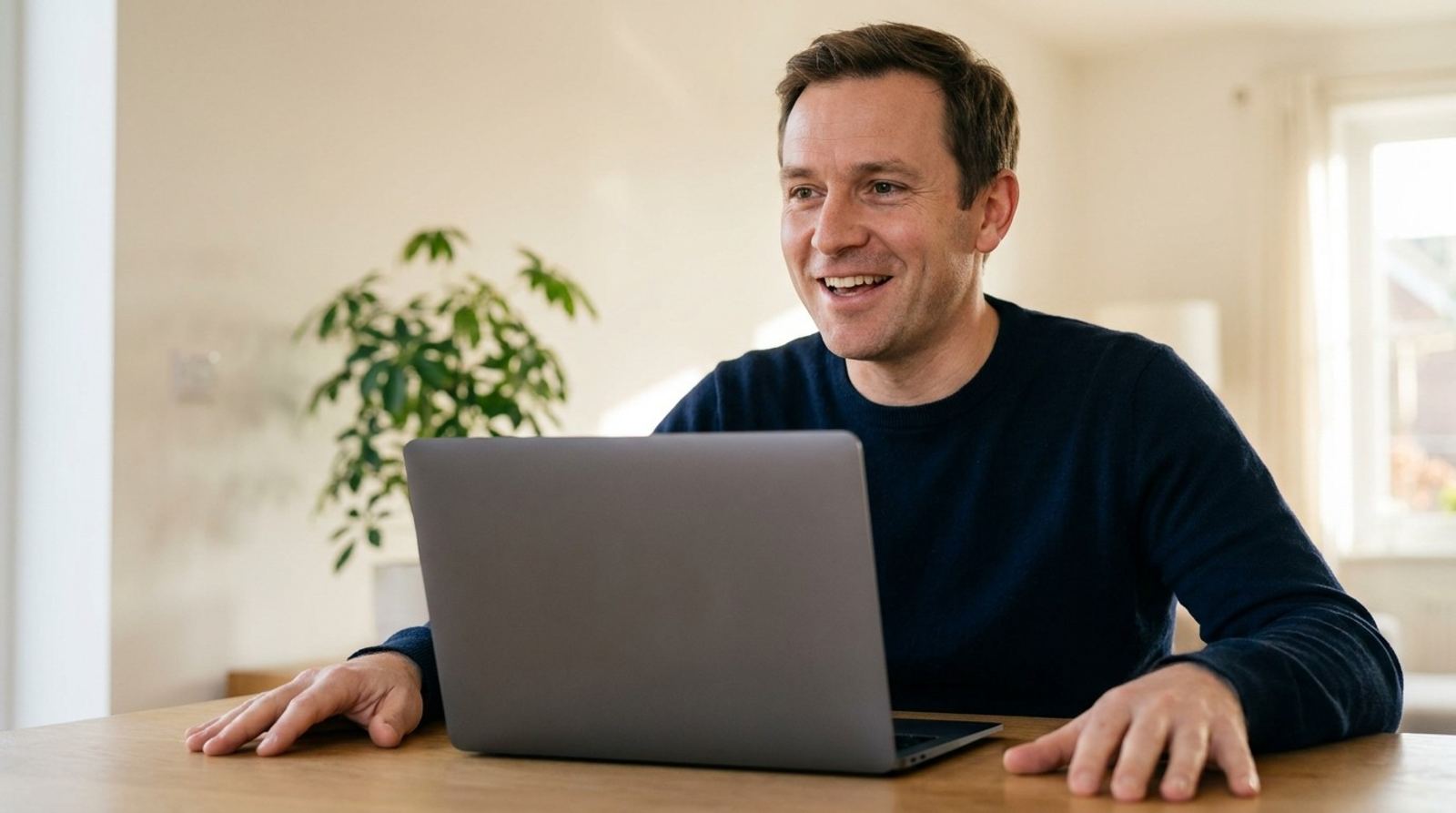 A man laughs in the middle of a video call at his living room table, both hands flat on either side of his laptop.
