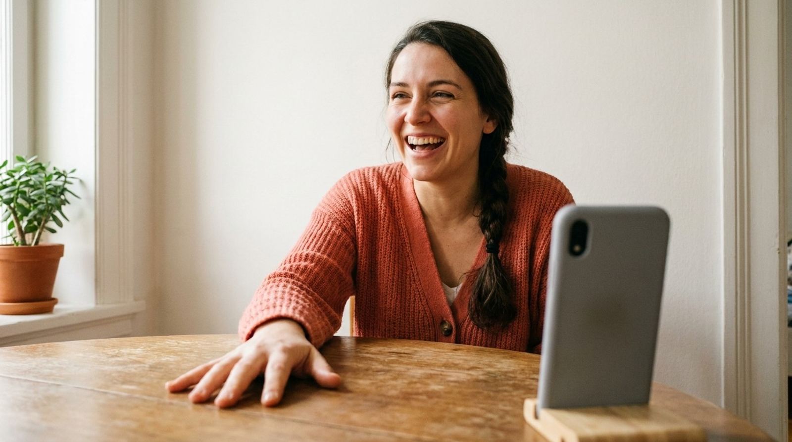 Une femme à la tresse rit en pleine conversation, la main posée sur la table, son téléphone dressé sur un support en bois.