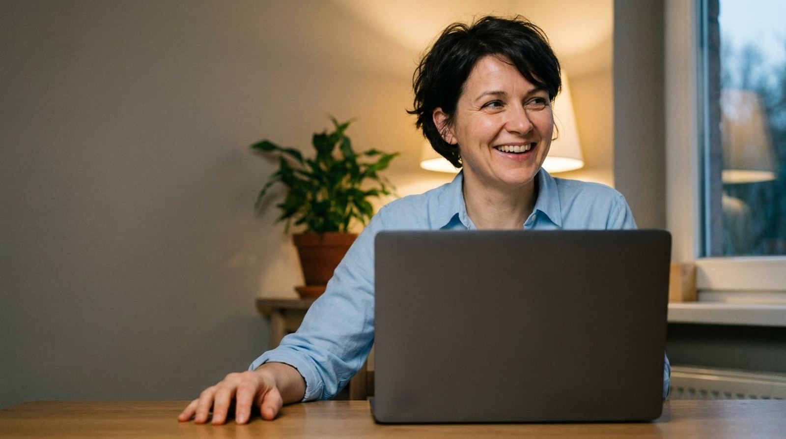 Une femme aux cheveux courts rit en parlant devant son ordinateur portable, la main posée sur la table, le soir près d’une lampe.