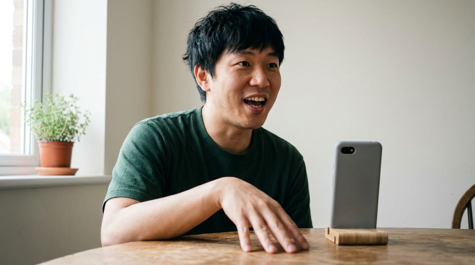 Un homme en t-shirt vert parle avec entrain à son téléphone posé sur un support en bois, la main sur la table.