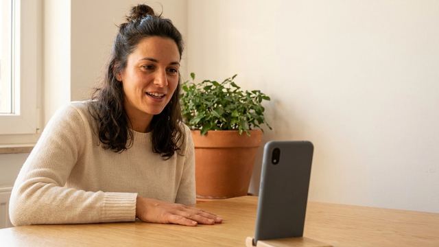 Une femme souriante, la main posée sur la table, parle à son téléphone installé sur un petit support, dans sa cuisine.