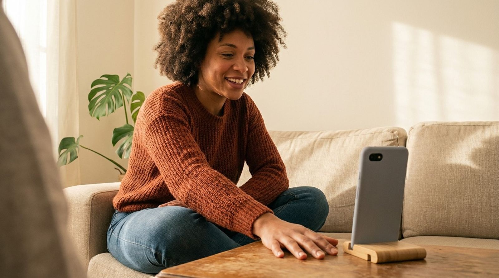 Une femme assise sur son canapé salue chaleureusement son partenaire d’échange, son téléphone dressé sur la table basse.