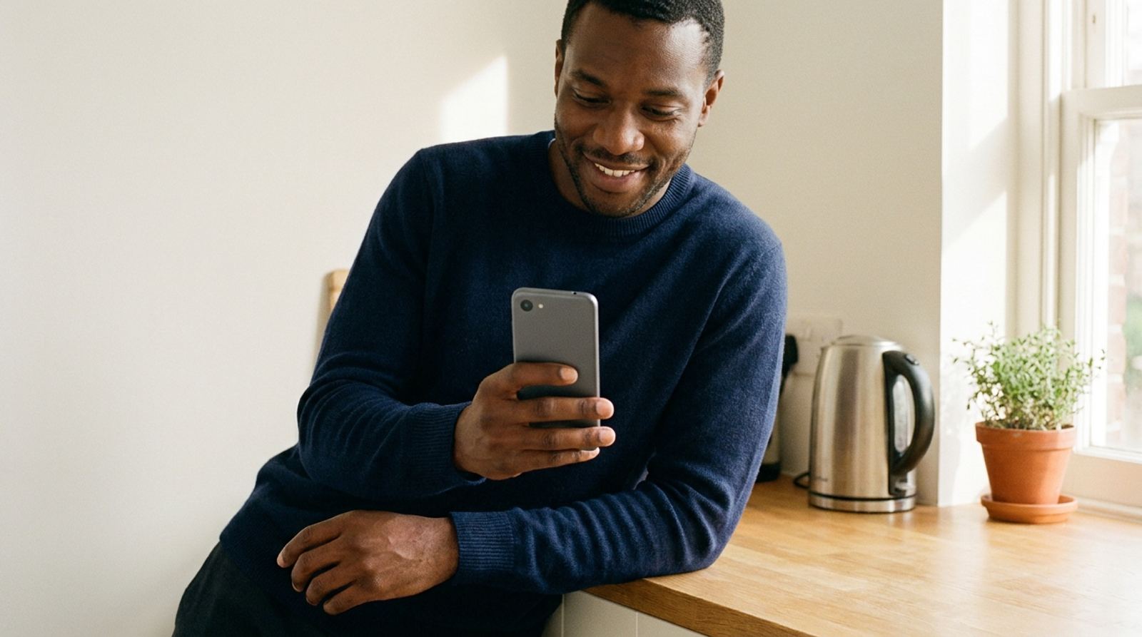 A man smiling at his phone, leaning against his kitchen counter.
