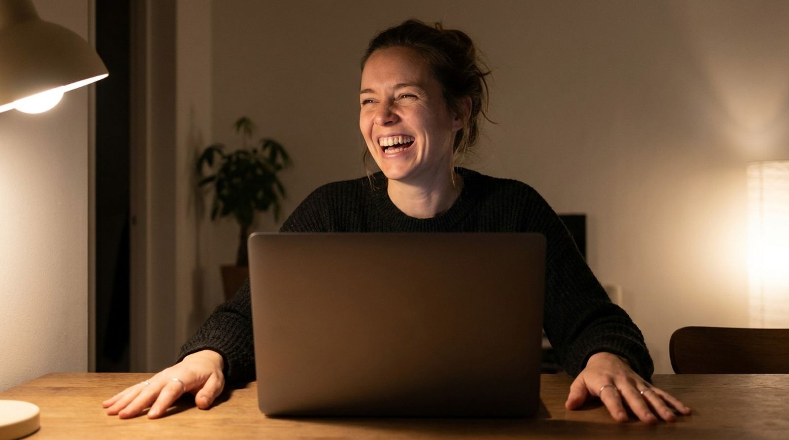 Une femme éclate de rire pendant un appel vidéo le soir, les mains posées de chaque côté de son ordinateur portable.