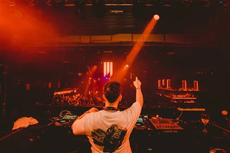 DJ with a raised fist facing the crowd under an orange spotlight