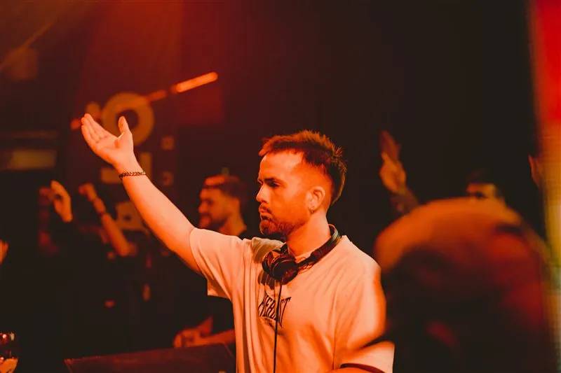 DJ with headphones around his neck raising an arm in a warm-lit club