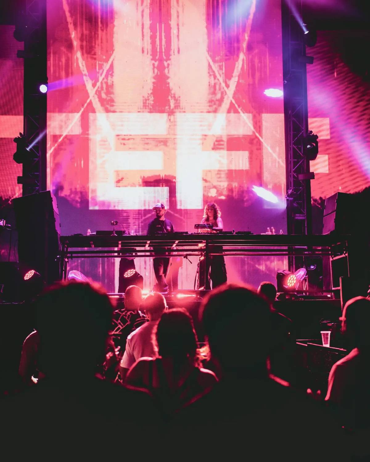 Two DJs on stage in front of a large pink and orange video screen, with the crowd in the foreground