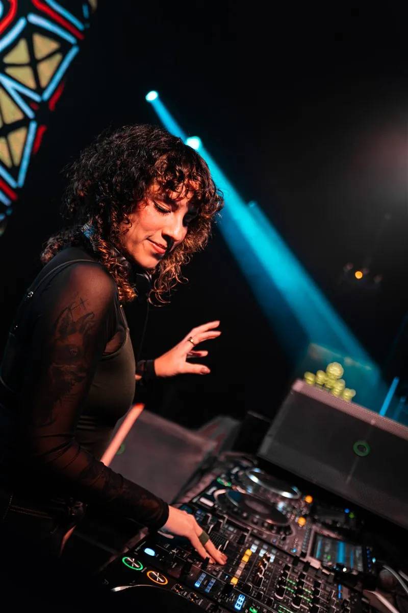 DJ with curly hair mixing on a controller in a dark club, with a blue beam of light behind