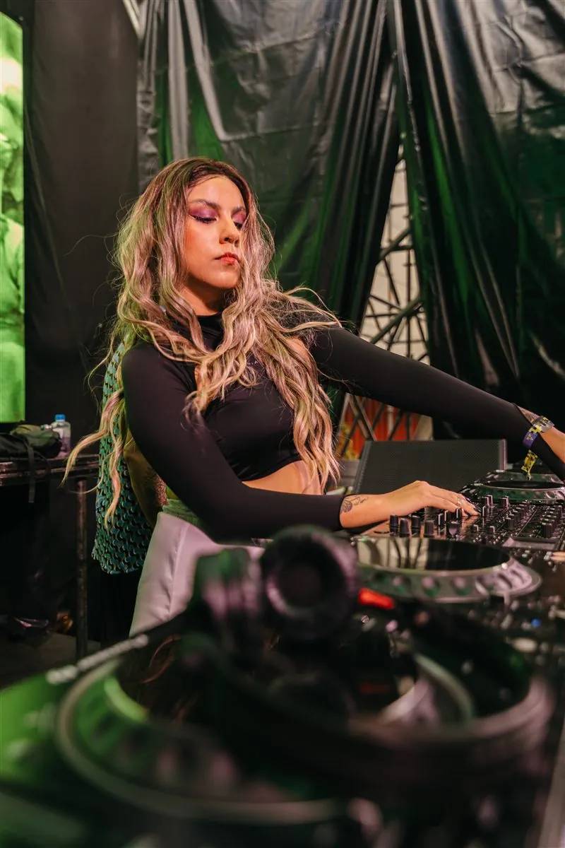 DJ with long wavy hair at the decks, with green curtains behind