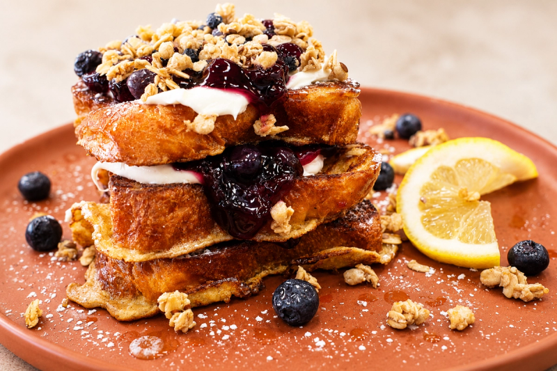 A stack of French toast topped with blueberry compote, cream and granola with a lemon slice