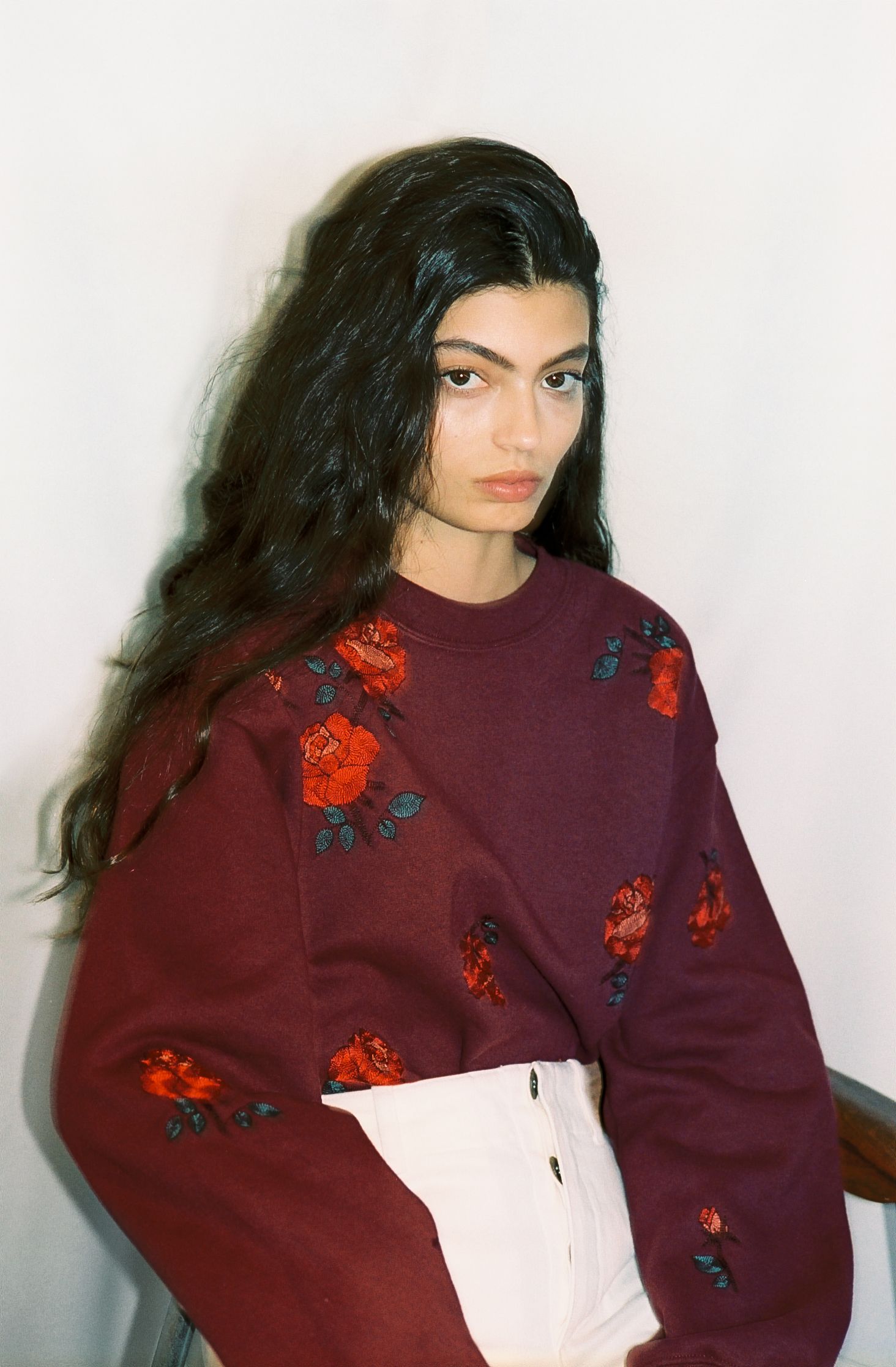 A young woman seated in a chair wearing a maroon sweater with a pattern of red embroidered roses