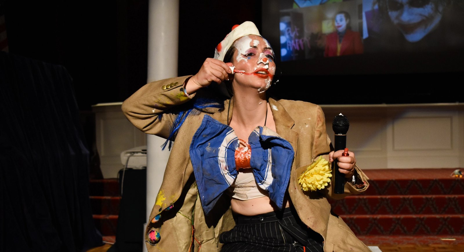 Alex Tatarsky in clown attire, with a mic in hand. They are messily applying makeup to their face.