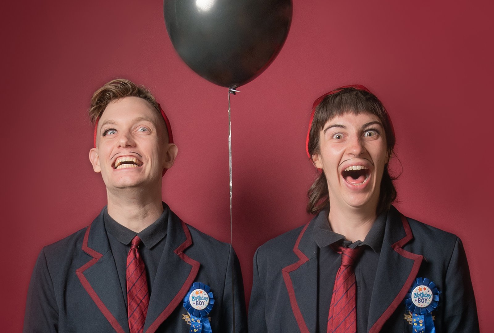 S.E. Grummet and Sam Kruger, in black suits with red ties stand in front of a red wall, holding a black balloon, and smiling widely at the camera.